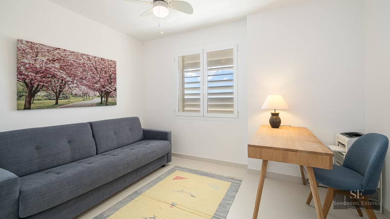 A minimalist room with a grey sofa, wooden desk with a lamp, a blue chair, and a window with white shutters.
