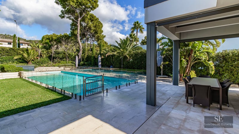 Piscine turquoise avec clôture en verre à côté d'une terrasse en pierre ombragée avec table et jardin tropical.