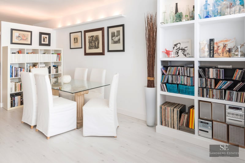 A bright dining room with a glass table, white chairs, and extensive white bookshelves filled with books and music.