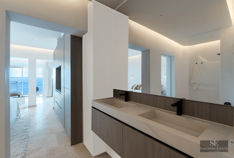 Modern master bathroom featuring double stone sinks, dark wood vanity, and a view through to a bedroom and the sea.