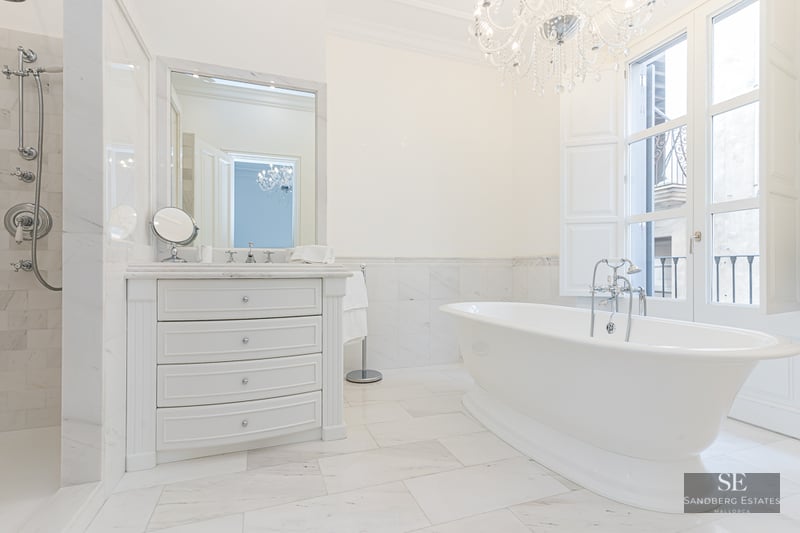 Luxury white marble bathroom featuring a freestanding tub, crystal chandelier, and large window with natural light.