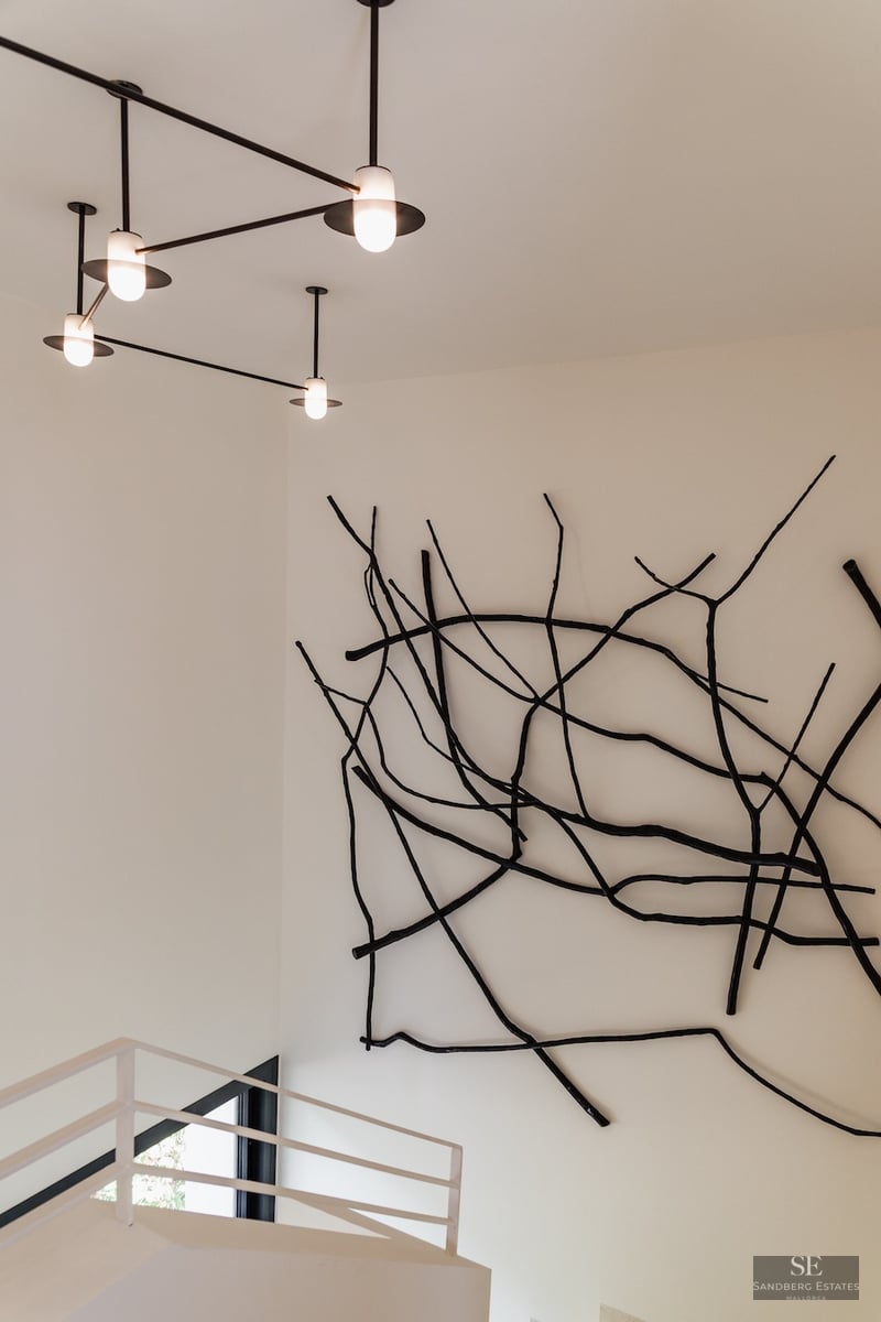 Black branch-like wall sculpture next to a modern geometric chandelier in a white minimalist room with stairs.