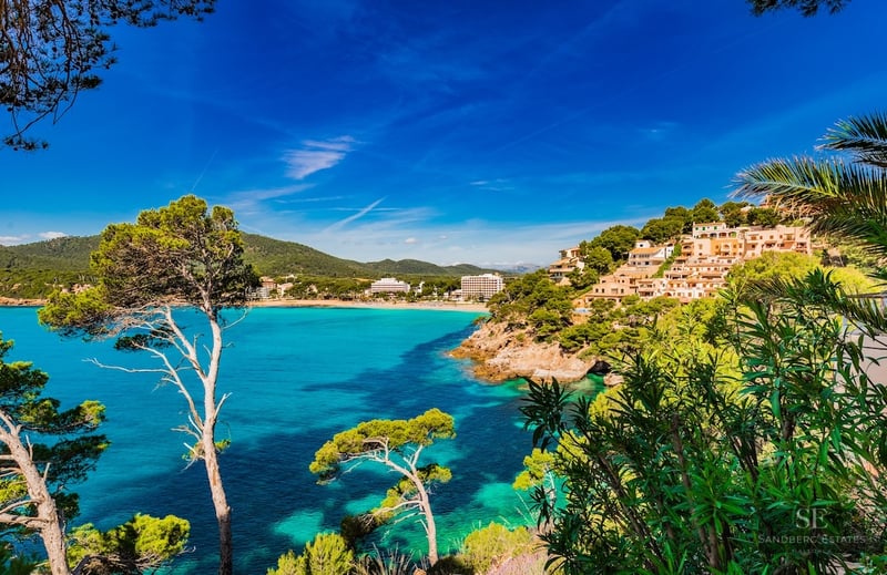 View of a coastal bay with turquoise waters, pine trees, and Mediterranean buildings under a blue sky.