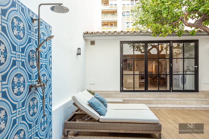 An outdoor shower with patterned blue tiles next to sun loungers on a wooden deck with a lemon tree.