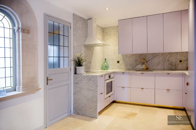Contemporary kitchen with white cabinets, marble backsplash, gold faucet, and an arched stone window.