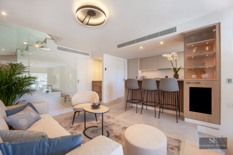 Modern living room with cream sofa, glass staircase, and a wood-paneled breakfast bar with grey stools.