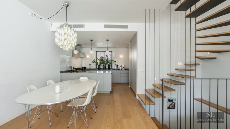 White dining room with oval table, Eames chairs, designer chandelier, grey kitchen, and wood/metal staircase.