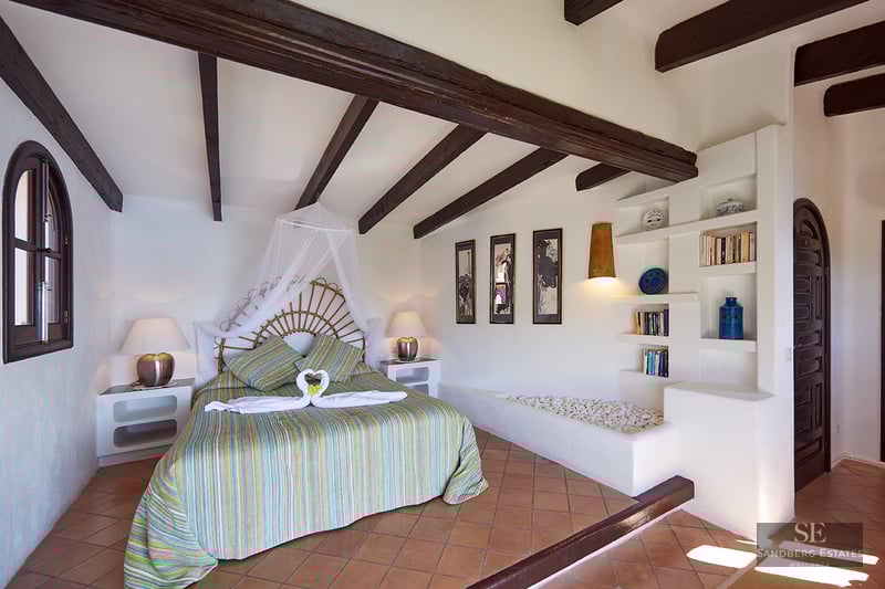 Bedroom with wicker headboard bed, dark wooden beams, terracotta floor, and white walls with built-in shelving.