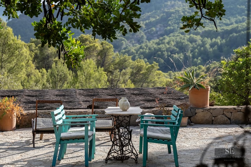Stone terrace with teal wooden chairs and a vintage table overlooking a lush green forest and mountain range.