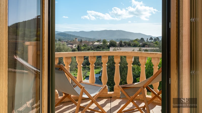 Sunlit balcony with stone balustrades, two wooden deck chairs, and a view of a village and distant mountains.