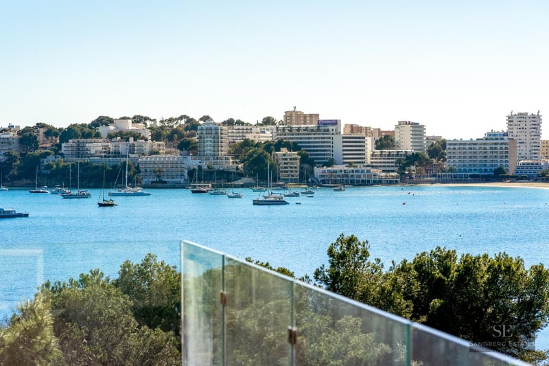 Glass balcony railing overlooking a bright blue bay filled with sailboats and a coastal city skyline.
