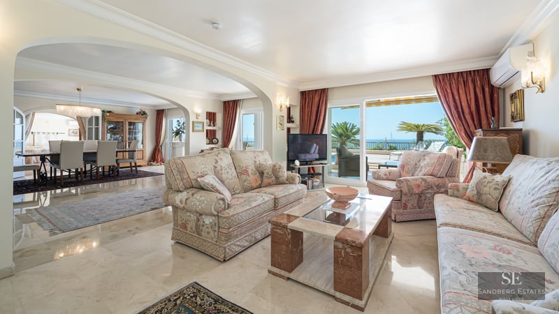 Bright living room with floral sofas, marble coffee table, and large glass doors opening to a terrace with sea view.