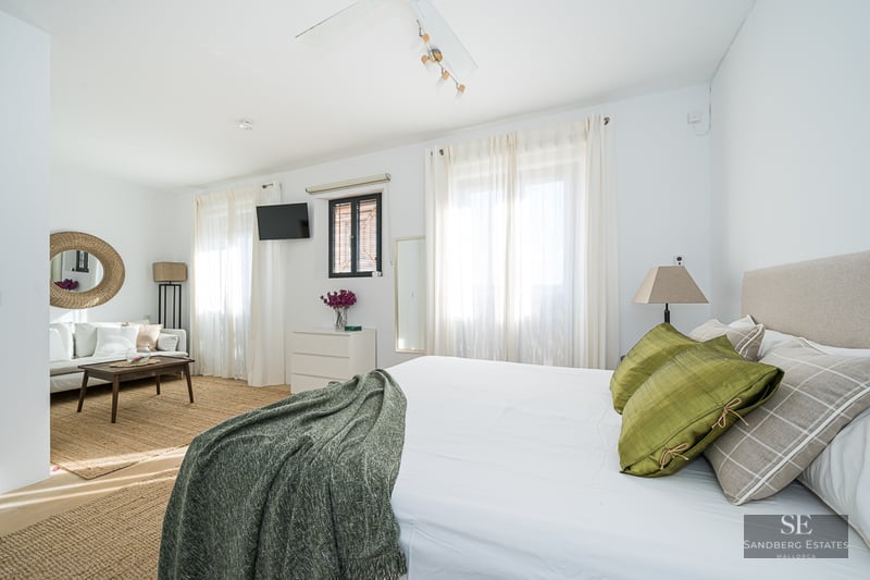 Bright master bedroom featuring a large bed with green pillows, a lounge area with a white sofa, and jute rugs.