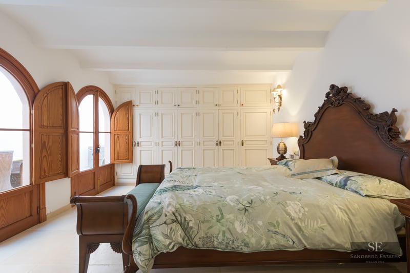 Spacious bedroom with a dark wood carved bed, floral bedding, white wardrobes, and arched wooden doors with shutters.