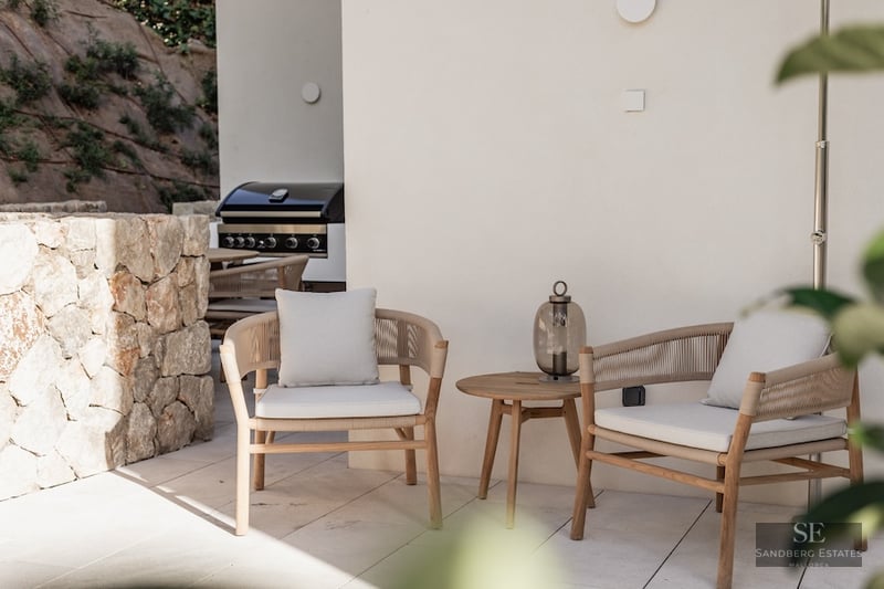 Two elegant modern armchairs with cushions next to a wooden side table on a stone terrace with a grill in the background.