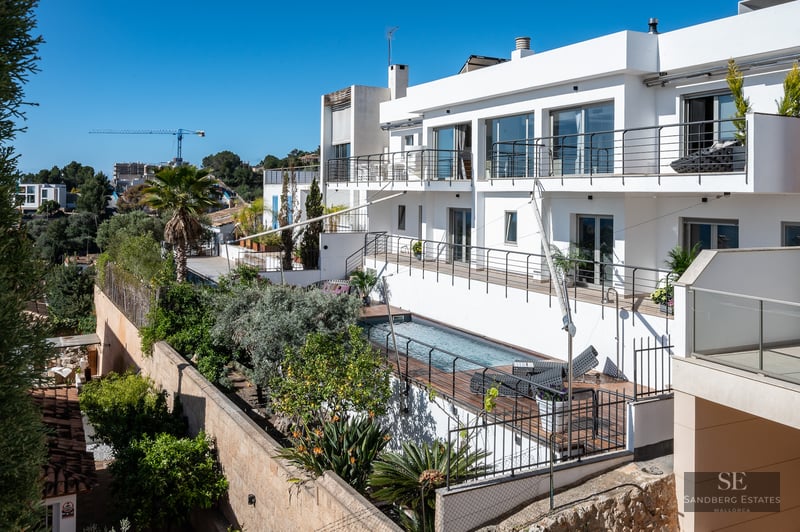 Modern white multi-level villa with a rectangular swimming pool, wooden decking, and lush Mediterranean landscaping.