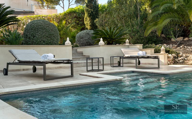 Blue swimming pool with modern sun loungers on a stone terrace surrounded by lush Mediterranean greenery.