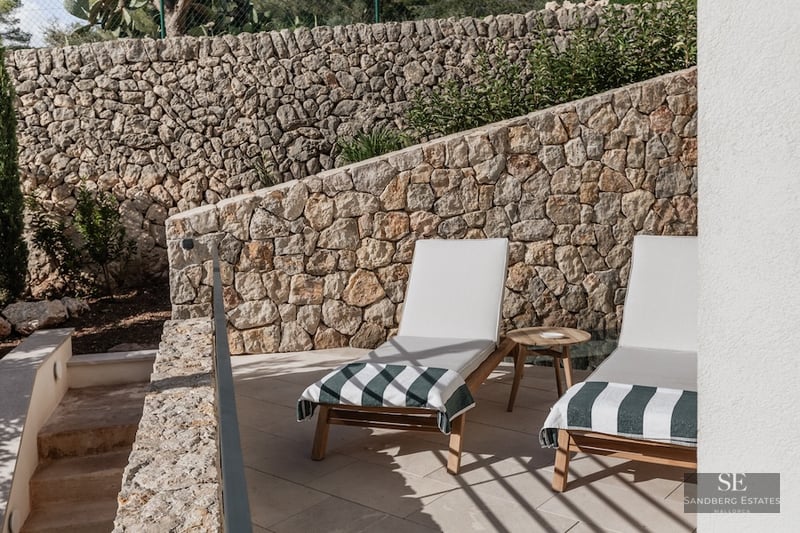 Two wooden sun loungers with white cushions in front of a rustic stone wall on a sunny terrace.