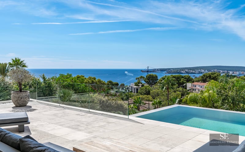Modern stone terrace featuring an infinity pool with a glass railing overlooking the blue sea and lush greenery.