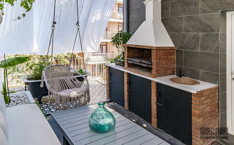 A modern terrace featuring a brick barbecue, a hanging macrame chair, a dining table, and decorative tiling.