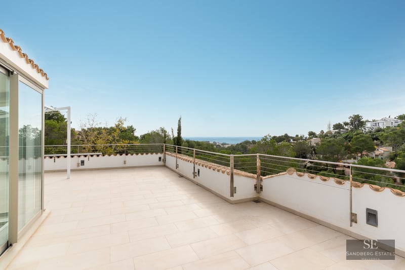 Large light-tiled terrace with a modern metal railing overlooking a lush green landscape and the sea.