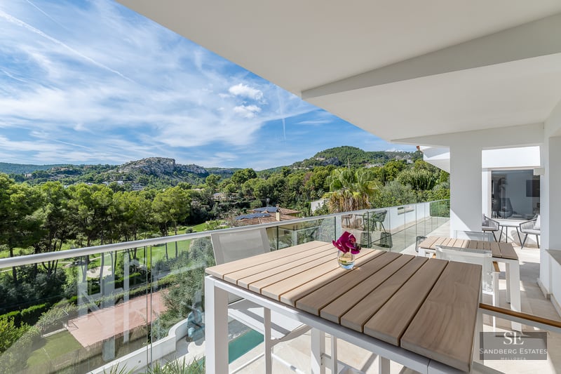 Modern balcony with wooden table and glass railing overlooking a lush green valley and mountains under a blue sky.