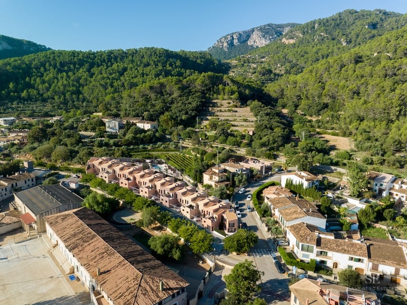 Aerial drone shot showing modern terracotta houses in a Mediterranean village surrounded by forested mountains.