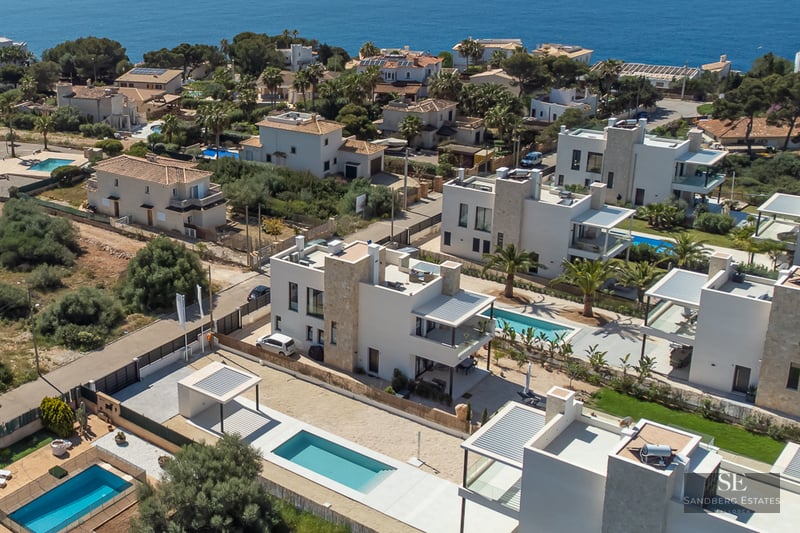 Drone view of modern white villas with private pools and stone accents in a coastal Mediterranean neighborhood.