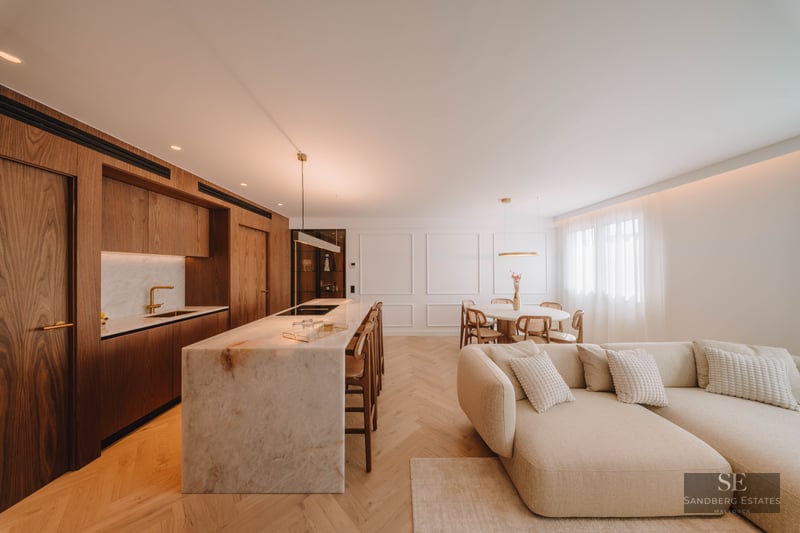 Modern open-plan room with a marble kitchen island, wood cabinets, herringbone floors, and a large cream sectional sofa.