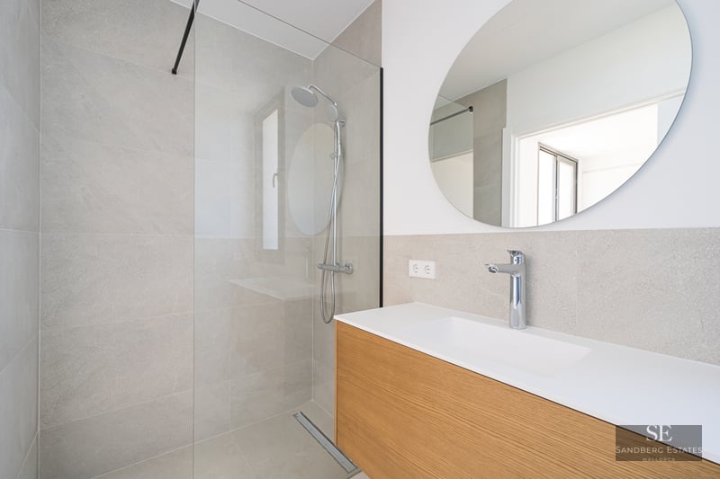 Modern bathroom with glass-enclosed shower, grey tiles, circular mirror, and wooden floating vanity.