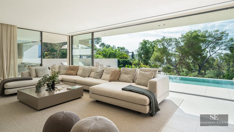 A bright, modern living room with a large cream sectional sofa looking out through glass walls to a private pool and trees.