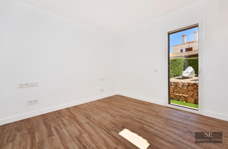 Empty room with white walls, wood floors, and a glass door opening to a garden with a stone wall.