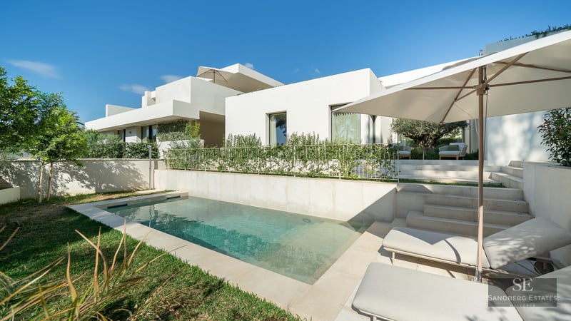 Modern white villa with a rectangular swimming pool, stone patio, sun loungers, and lush green landscaping under a blue sky.