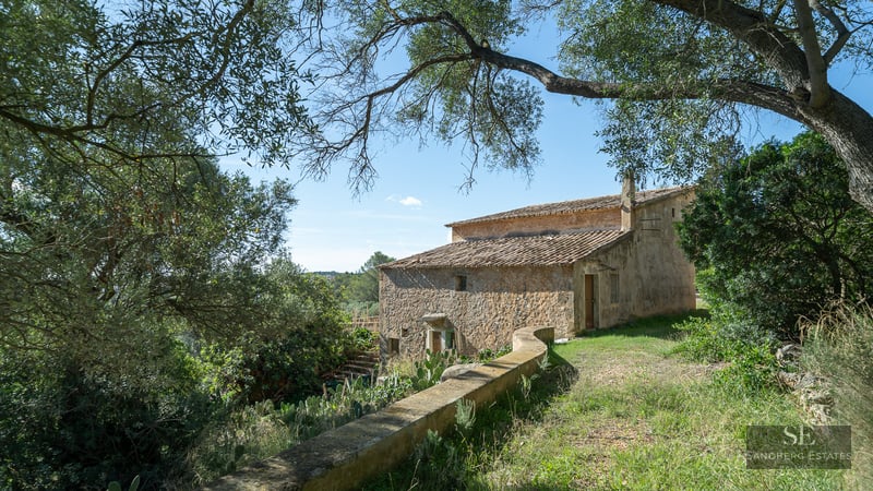 A historic two-story stone house with a terracotta roof nestled among trees under a clear blue sky.