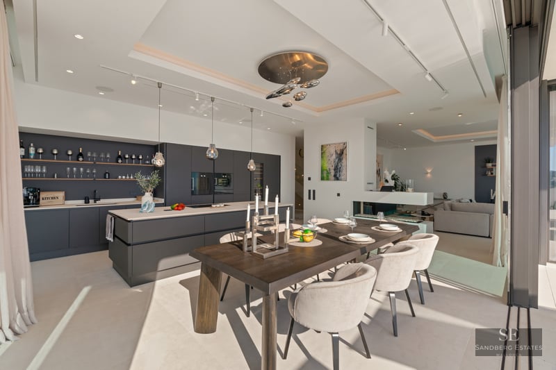 Modern dining area with dark wood table, beige chairs, and sleek gray kitchen featuring floor-to-ceiling windows.