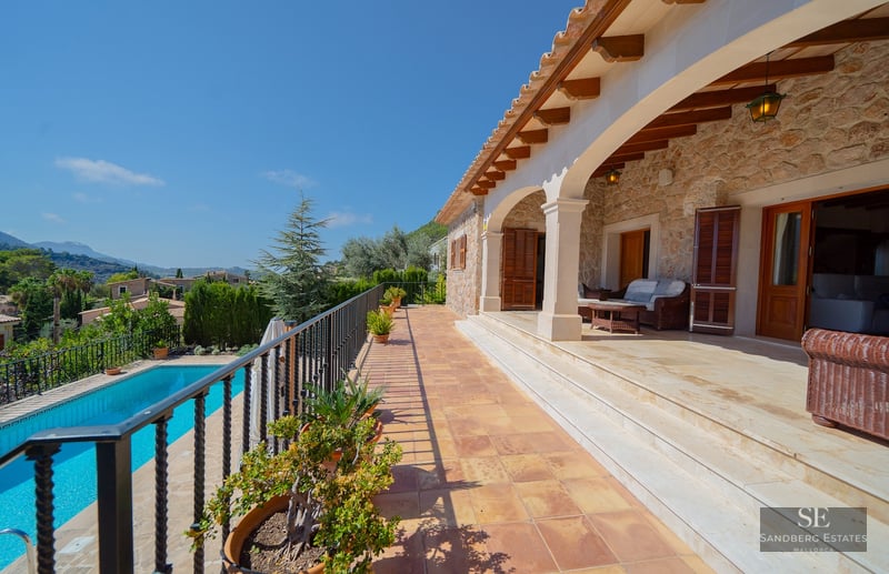 Shaded stone terrace with wooden beams, iron railing, and views of a blue swimming pool and distant mountains.