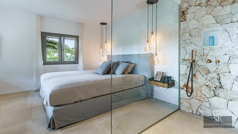 Contemporary bedroom with a grey bed, glass wall divider, and a rustic stone shower with brass fixtures.