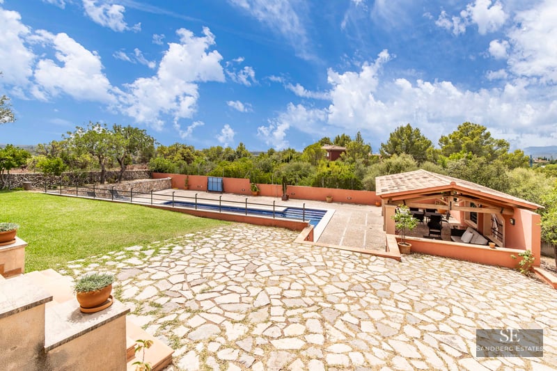 Large stone patio with swimming pool, green lawn, and a covered lounge area with wooden beams and tiled roof.