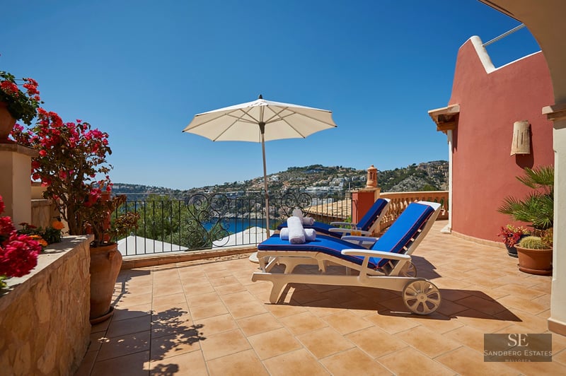 Sunny terrace with two blue lounge chairs, white umbrella, and panoramic sea and coastal hillside views.