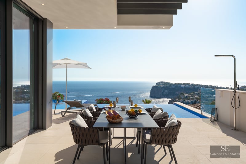 Modern outdoor terrace with a dining table overlooking an infinity pool and a vast blue ocean with coastal cliffs.