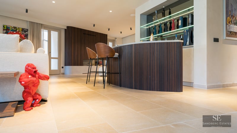 Modern living room featuring a curved wooden bar, designer stools, stone floors, and a bright red gorilla sculpture.