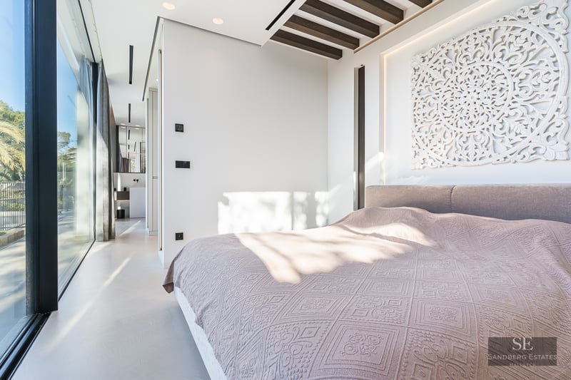 Bright modern bedroom with a large bed, floor-to-ceiling glass doors, and a decorative white carved headboard.