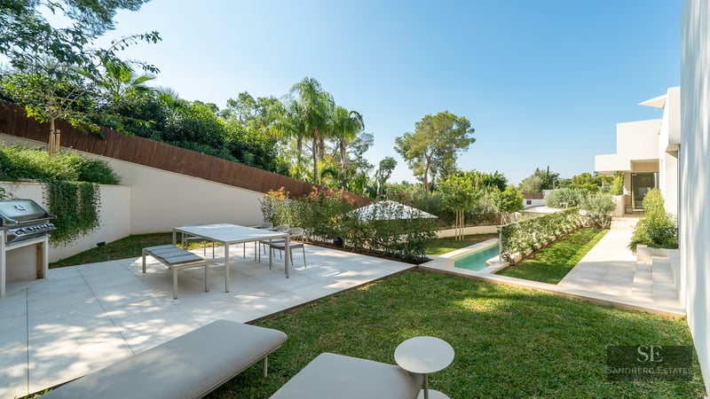 Modern white terrace featuring a dining area, outdoor grill, and lap pool surrounded by lush landscaping and palm trees.