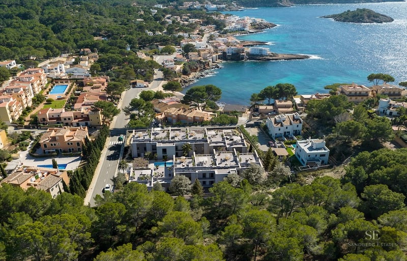 Drone view of a modern gray apartment complex nestled between pine forests and a turquoise Mediterranean bay.
