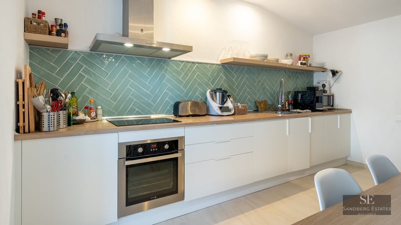 Modern white kitchen featuring a teal herringbone tile backsplash, wooden countertops, and stainless steel appliances.