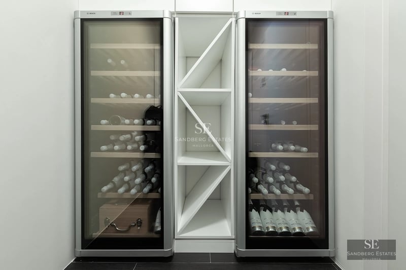 Two tall glass-front wine coolers with wooden shelves flanking a white geometric storage unit on a dark tile floor.