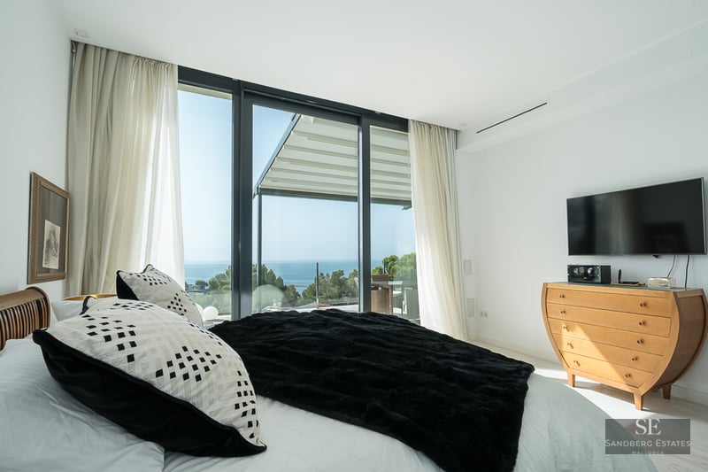 Modern bedroom with large bed, black throw, and floor-to-ceiling glass doors opening to a stunning sea view.