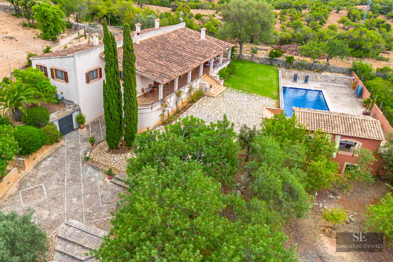 High-angle view of a villa with a terracotta roof, stone courtyard, blue swimming pool, and lush green landscaping.