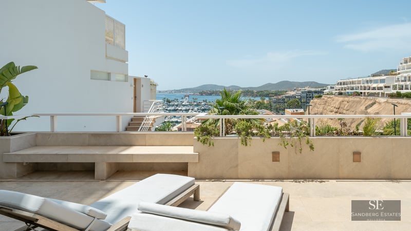 Sun loungers on a modern stone terrace overlooking a marina with boats and mountains under a clear blue sky.
