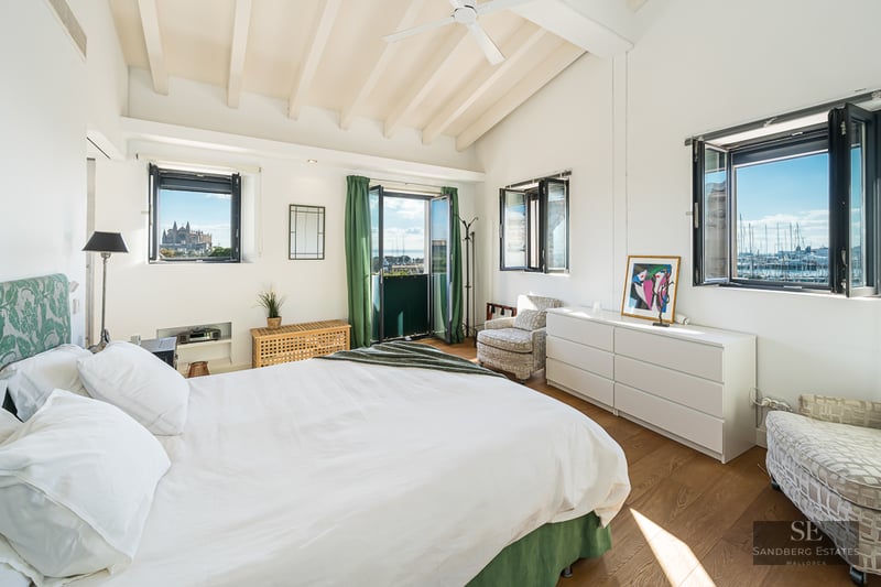 Bright bedroom with white walls, wooden beams, white bed, and windows overlooking a harbor and cathedral.